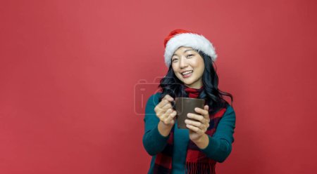 Photo for Pretty smiling asian woman in warm christmas sweater and santa hat is having hot chocolate on red background for season celebration - Royalty Free Image