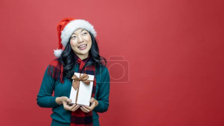 Téléchargez les photos : Jolie femme asiatique souriante en pull de Noël chaud et chapeau santa tenant boîte cadeau comme cadeau avec fond rouge pour la célébration de la saison - en image libre de droit