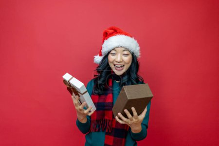 Téléchargez les photos : Jolie femme asiatique souriante en chandail de Noël chaud et chapeau de Père Noël boîte cadeau de déballage comme présent avec fond rouge le jour de la boxe pour l'utilisation de la célébration de la saison - en image libre de droit