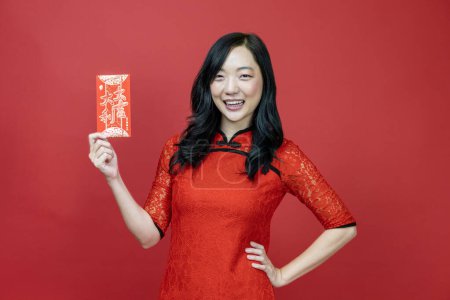 Téléchargez les photos : Femme asiatique tenant enveloppe fortune argent rouge bénédiction mot chinois qui signifie "Puissiez-vous avoir une grande chance et un grand profit" isolé sur fond rouge pour la célébration du Nouvel An chinois - en image libre de droit