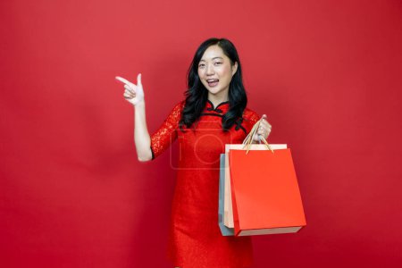 Photo for Asian chinese woman with red cheongsam or qipao holding shopping bag wish the good luck sale and prosperity in Chinese New Year celebration holiday isolated on red background concept - Royalty Free Image