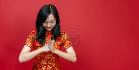 Photo for Asian chinese woman with red cheongsam or qipao doing polite respectful gesture for wishing the good luck and prosperity in Chinese New Year celebration holiday isolated on red background usage - Royalty Free Image