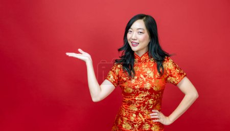 Téléchargez les photos : Femme asiatique avec cheongsam rouge ou qipao pointant pour présenter pour la fête du Nouvel An chinois vacances isolées sur fond rouge - en image libre de droit