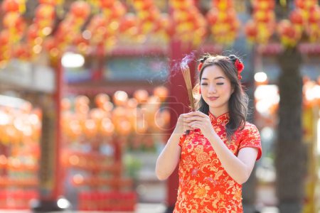 Foto de Asian woman in red cheongsam qipao dress is offering incense to the ancestral god inside Chinese Buddhist temple during lunar new year for  for best wish blessing and good luck - Imagen libre de derechos