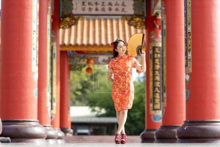 Téléchargez les photos : Asian woman in red cheongsam qipao dress holding paper fan while visiting the Chinese Buddhist temple during lunar new year for traditional culture - en image libre de droit