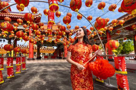 Téléchargez les photos : Asian woman in red cheongsam qipao dress holding lantern while visiting the Chinese Buddhist temple during lunar new year for traditional culture - en image libre de droit