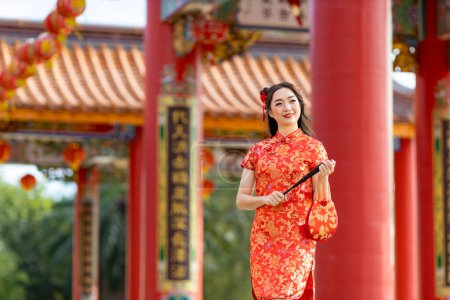 Téléchargez les photos : Asian woman in red cheongsam qipao dress holding paper fan while visiting the Chinese Buddhist temple during lunar new year for traditional culture - en image libre de droit
