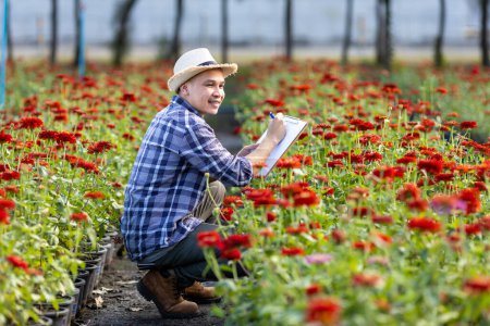 Téléchargez les photos : Asian gardener is taking note using clip board on the growth and health of red zinnia plant while working in his rural field farm for medicinal herb and cut flower - en image libre de droit