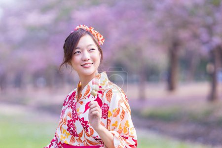 Foto de Mujer japonesa con vestido tradicional de kimono sosteniendo dulce postre de hanami dango mientras camina en el parque en el árbol de flor de cerezo durante el festival de sakura de primavera con espacio para copiar - Imagen libre de derechos