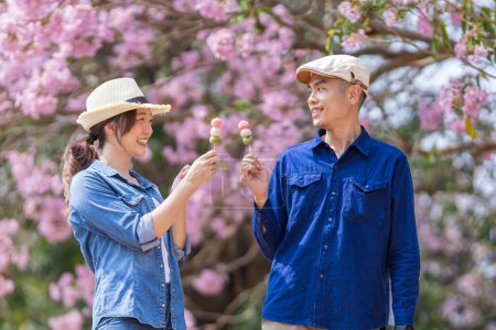 Foto de Joven pareja asiática celebrando el dulce postre de hanami dango mientras camina en el parque en el árbol de flor de cerezo durante el festival de sakura de primavera - Imagen libre de derechos