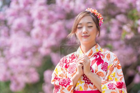 Téléchargez les photos : Femme japonaise dans la robe traditionnelle kimono fait un voeu de bonne année pour la bonne fortune tout en marchant dans le parc au cerisier pendant le festival sakura printemps - en image libre de droit