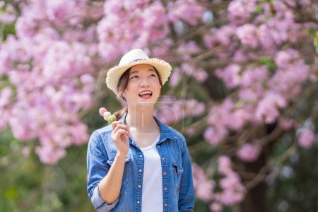 Téléchargez les photos : Femme asiatique tenant le doux dessert hanami dango tout en marchant dans le parc au cerisier pendant le festival de sakura de printemps - en image libre de droit