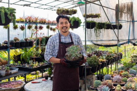 Foto de Jardinero asiático está trabajando dentro del invernadero lleno de plantas suculentas colección mientras sostiene una olla saludable para jardín ornamental y ocio hobby - Imagen libre de derechos