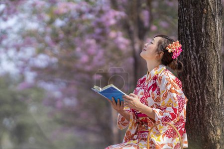 Téléchargez les photos : Femme japonaise en robe kimono traditionnelle assise sous un cerisier en fleurs tout en lisant un livre pendant le festival de sakura de printemps - en image libre de droit