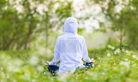 Téléchargez les photos : Vue arrière de la femme en sweat à capuche pratique relaxant yoga méditation dans la forêt pleine de fleurs de marguerite en été pour atteindre le bonheur de la paix intérieure sagesse avec la lumière du matin pour un esprit sain - en image libre de droit
