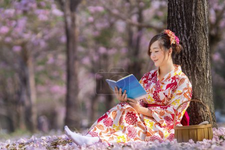 Photo for Japanese woman in traditional kimono dress sitting under cherry blossom tree while reading a book during spring sakura festival - Royalty Free Image