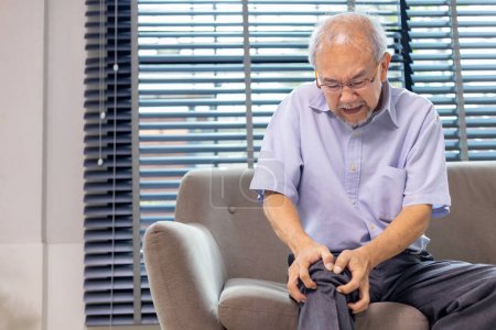 Foto de Hombre asiático mayor que sufre de osteoartritis de rodilla síntoma whiling sentado en el sofá en casa con espacio de copia para el tratamiento de la cirugía médica y la terapia física - Imagen libre de derechos