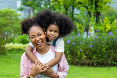 Téléchargez les photos : African American mother is playing piggyback riding and hugging with her young daughter while having a summer picnic in the public park for wellbeing and happiness - en image libre de droit