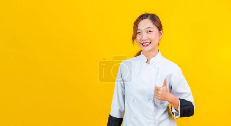 Foto de Retrato estudio disparo de mujer asiática chef con uniforme profesional posando con los pulgares hacia arriba en confidente aislado sobre fondo amarillo para culinaria y restaurante concepto - Imagen libre de derechos