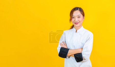 Foto de Retrato estudio disparo de mujer asiática chef con uniforme profesional posando con la mano cruzada en confidente aislado sobre fondo amarillo para culinaria y restaurante concepto - Imagen libre de derechos