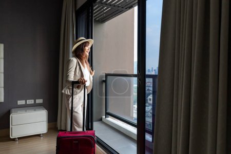 Téléchargez les photos : Femme asiatique déplaçant l'hôtel de luxe avec bagages en vacances tout en regardant la vue de la fenêtre avec immeuble de grande hauteur dans le centre-ville urbain pour le voyage d'affaires et l'utilisation de la copropriété - en image libre de droit