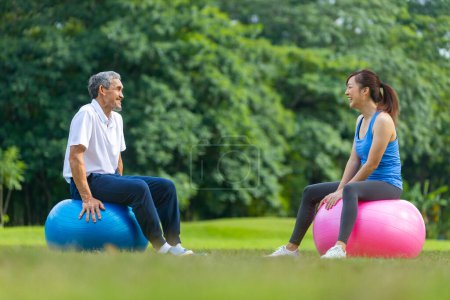Téléchargez les photos : Senior homme asiatique et sa fille est assis sur la balle de yoga dans le parc public tout en ayant conversation de loisirs ensemble pour l'exercice de longévité aîné et l'entraînement en plein air - en image libre de droit