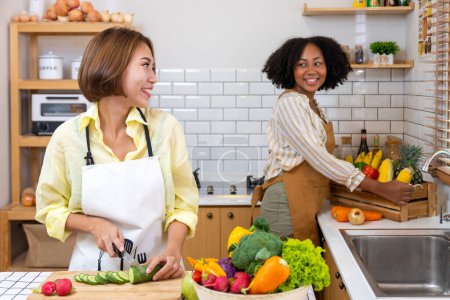 Foto de Mujer asiática y su amiga afroamericana se están ayudando mutuamente a preparar alimentos saludables con variedad de vegetales mientras lavan y cortan el ingrediente - Imagen libre de derechos