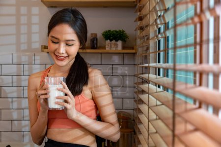 Foto de Mujer asiática saludable bebiendo un vaso de leche para mientras usa ropa deportiva para la nutrición y suplemento de proteína en la ventana para el embarazo y el beneficio de maternidad - Imagen libre de derechos