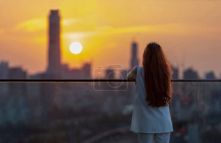 Photo for Woman looking and enjoying sunset view from balcony with the sun setting behind skyscraper in busy urban downtown with loneliness for solitude, loneliness and dreaming of freedom lifestyle - Royalty Free Image