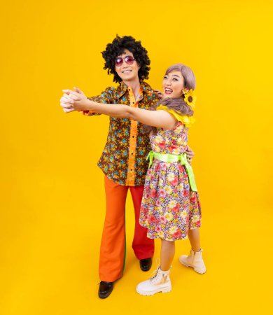Téléchargez les photos : Robe de couple hippie asiatique dans les années 80 mode vintage avec des vêtements rétro colorés tout en dansant ensemble isolé sur fond jaune pour une soirée déguisée et la culture pop - en image libre de droit