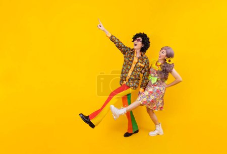 Téléchargez les photos : Robe de couple hippie asiatique dans les années 80 mode vintage avec des vêtements rétro colorés tout en dansant ensemble isolé sur fond jaune pour une soirée déguisée et la culture pop - en image libre de droit