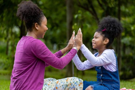 Photo for African American mother is laughing while playing patty cake with her young daughter while having a summer picnic in the public park for wellbeing and happiness - Royalty Free Image