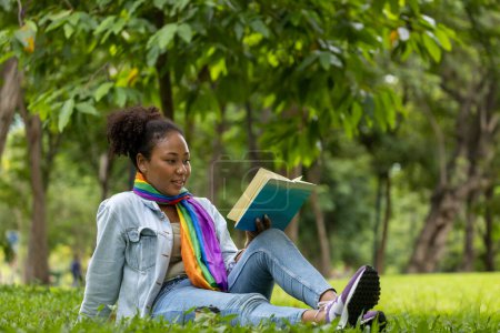 Photo for African American woman is lying down in the grass lawn inside the public park holding book in her hand during summer for reading and education - Royalty Free Image