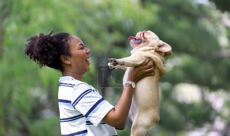 Foto de Mujer afroamericana está jugando con su cachorro bulldog francés mientras pasea por el parque de perros en el césped de hierba después de hacer ejercicio matutino durante el verano - Imagen libre de derechos