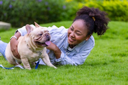 Photo for African American woman is playing with her french bulldog puppy while lying down in the grass lawn after having morning exercise in public park - Royalty Free Image
