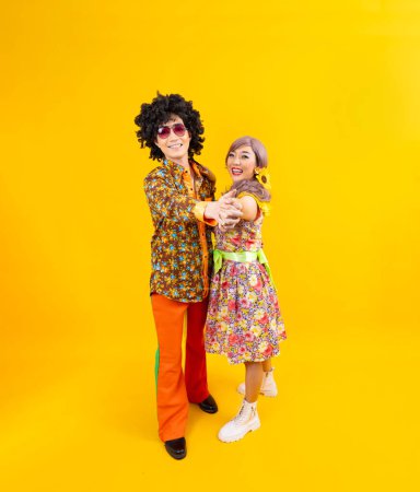 Foto de Vestido de pareja hippie asiático en 80 vintage colorido moda retro ropa mientras bailan juntos aislados en el fondo amarillo para la fiesta de disfraces de lujo y la cultura pop - Imagen libre de derechos
