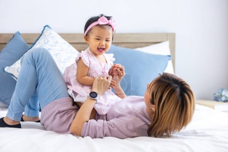 Téléchargez les photos : Mère asiatique joue avec sa jolie fille bébé souriante tout en passant du temps de qualité dans le lit pour le bonheur de la famille et la parentalité - en image libre de droit