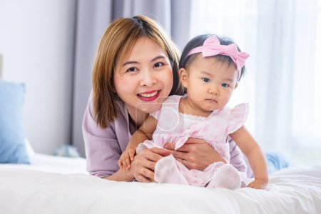 Téléchargez les photos : Mère asiatique tient sa jolie fille bébé souriante tout en passant du temps de qualité au lit pour le bonheur de la famille et le concept parental - en image libre de droit