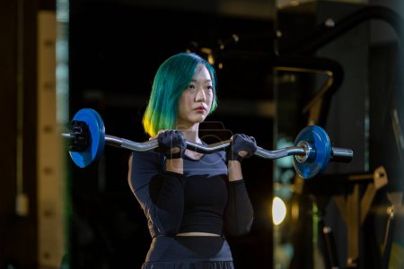 Foto de Mujer asiática es la práctica de levantamiento de pesas utilizando barra fácil como principiante en la barra para el brazo y el músculo del núcleo dentro del gimnasio con fondo oscuro para el ejercicio y el entrenamiento - Imagen libre de derechos