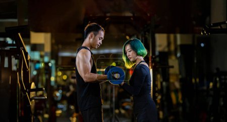 Téléchargez les photos : Femme asiatique utilise barre facile comme débutant dans la formation de poids sur haltère pour le muscle du bras à l'intérieur de la salle de gym avec le soutien de l'entraîneur pour prévenir les blessures fond sombre pour l'exercice et l'entraînement - en image libre de droit