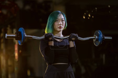 Photo for Asian woman is practice weight lifing using easy bar as beginner on barbell for arm and core muscle inside gym with dark background for exercising and workout - Royalty Free Image