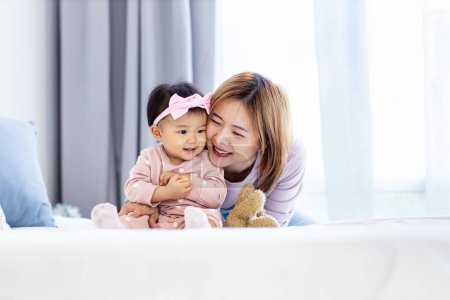 Téléchargez les photos : Mère asiatique tient sa jolie fille bébé souriante tout en passant du temps de qualité dans le lit pour le bonheur de la famille et la parentalité - en image libre de droit