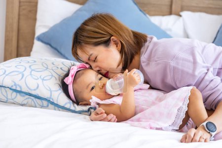 Téléchargez les photos : Mère asiatique nourrit le lait de la bouteille à son mignon tout-petit bébé fille au lit pour la nutrition saine des enfants et la maternité en élevant l'enfant à grandir et le concept parental - en image libre de droit