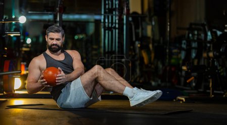 Foto de Ejercicio de hombre caucásico en gimnasio haciendo ejercicio de giro oblicuo sentado con bola de medicina de peso en el suelo que fortalece los abdominales y el músculo corporal central - Imagen libre de derechos