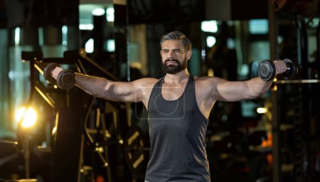Téléchargez les photos : Homme de sport musculaire barbe caucasienne pratique la musculation sur haltères doubles pour biceps et triceps muscle à l'intérieur de la salle de gym avec fond sombre pour l'exercice et l'entraînement - en image libre de droit