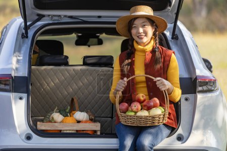 Téléchargez les photos : Heureuse agricultrice asiatique portant récolte de fruits avec des produits biologiques maison pomme, courge et citrouille assis sur le coffre de la voiture au marché agricole local pendant la saison d'automne pour les produits agricoles - en image libre de droit