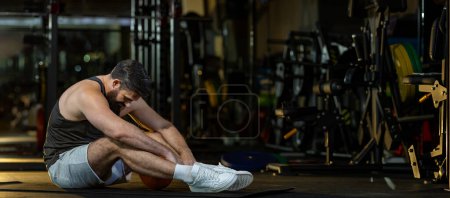 Foto de Hombre caucásico se enfrenta al síndrome de sobreentrenamiento después del entrenamiento dentro del gimnasio con fondo oscuro para el ejercicio y la aptitud - Imagen libre de derechos