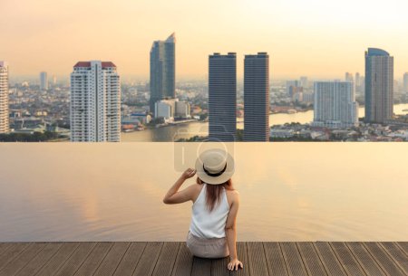 Photo for Pretty tourist woman is relaxing and enjoying her rooftop swimming pool view with cityscape background for vacation and travel at the sunset with skyscraper and downtown highrise condominium - Royalty Free Image