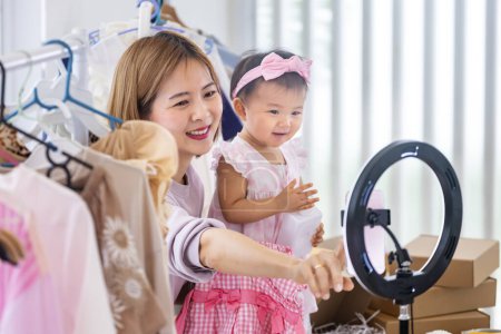 Téléchargez les photos : Jeune asiatique influenceur mère vend des vêtements pour enfants en direct en streaming à la maison tout en élevant son tout-petit comme assistance - en image libre de droit