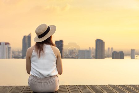 Photo for Pretty tourist woman is relaxing and enjoying her rooftop swimming pool view with cityscape background for vacation and travel at sunset with skyscraper and downtown highrise condominium - Royalty Free Image